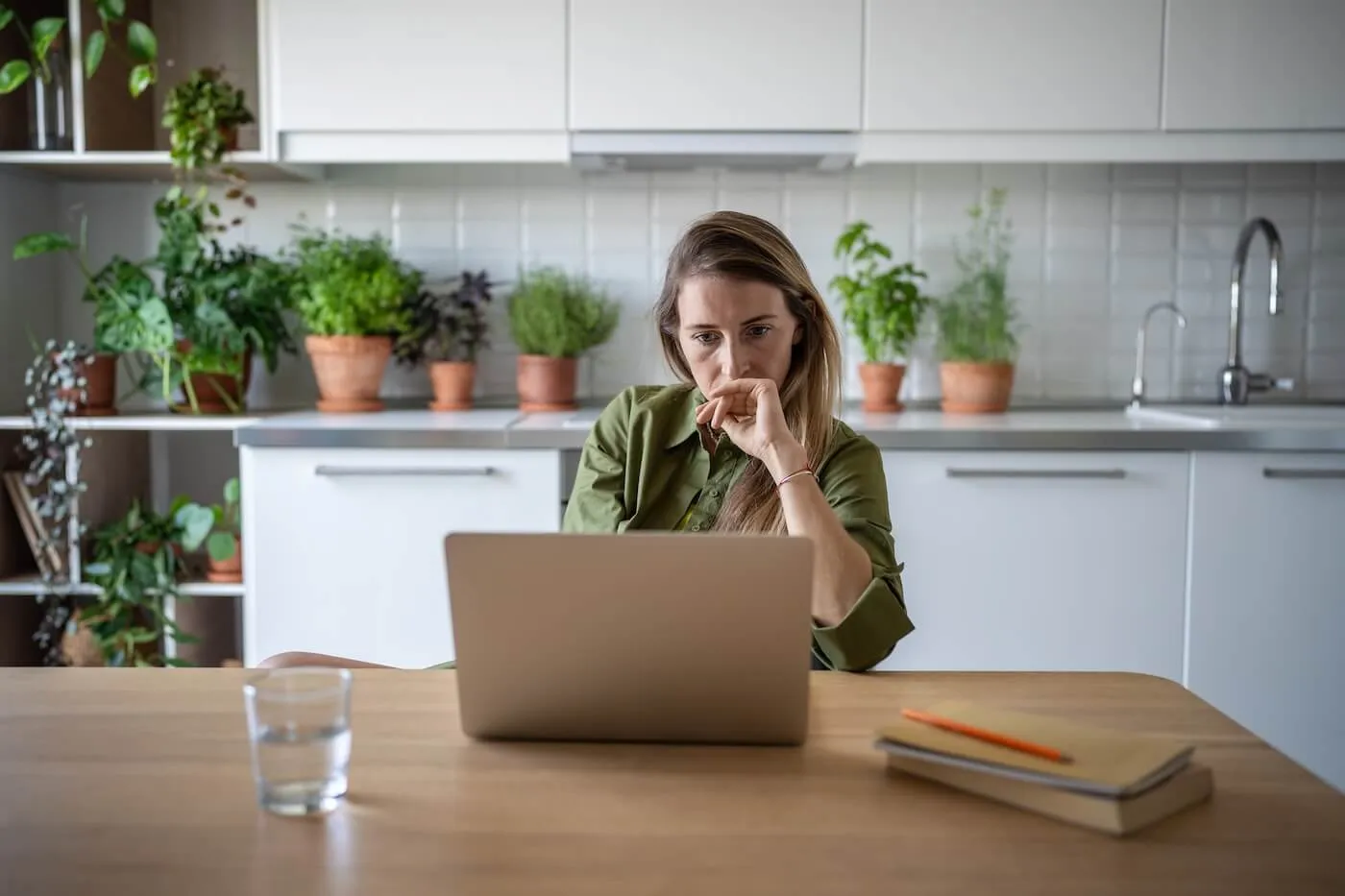 Focused woman seated at a kitchen table using a laptop, with one hand resting near the chin while looking at the screen. A notebook, pencil, and glass of water sit on the table, while potted plants, white cabinetry, and a modern kitchen are visible in the background.