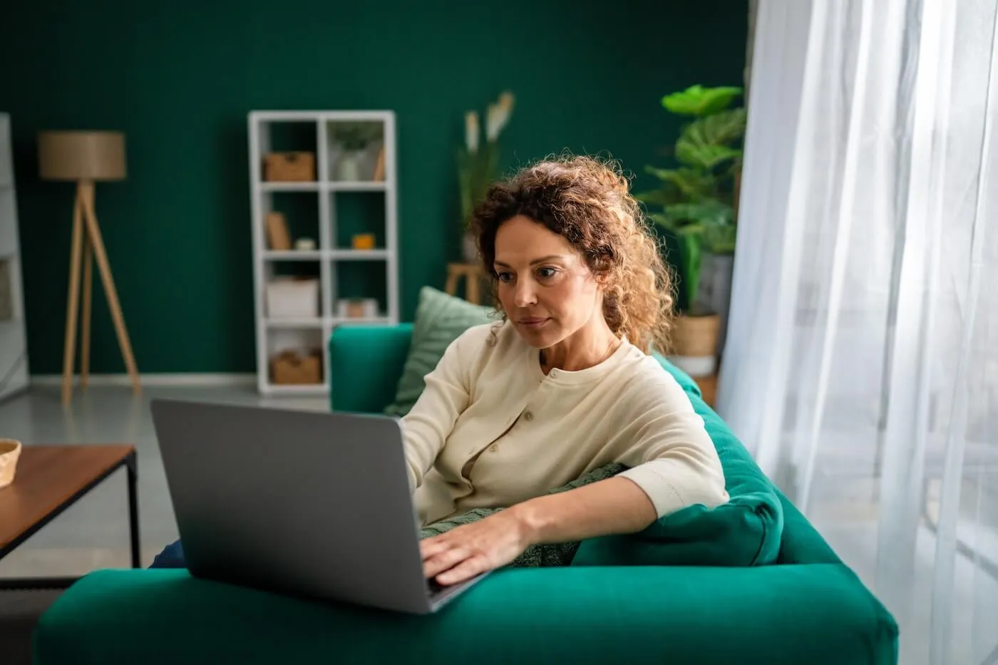 Focused woman sitting on a green sofa using a laptop in a modern living room, with plants, shelving, and large windows providing natural light in the background.