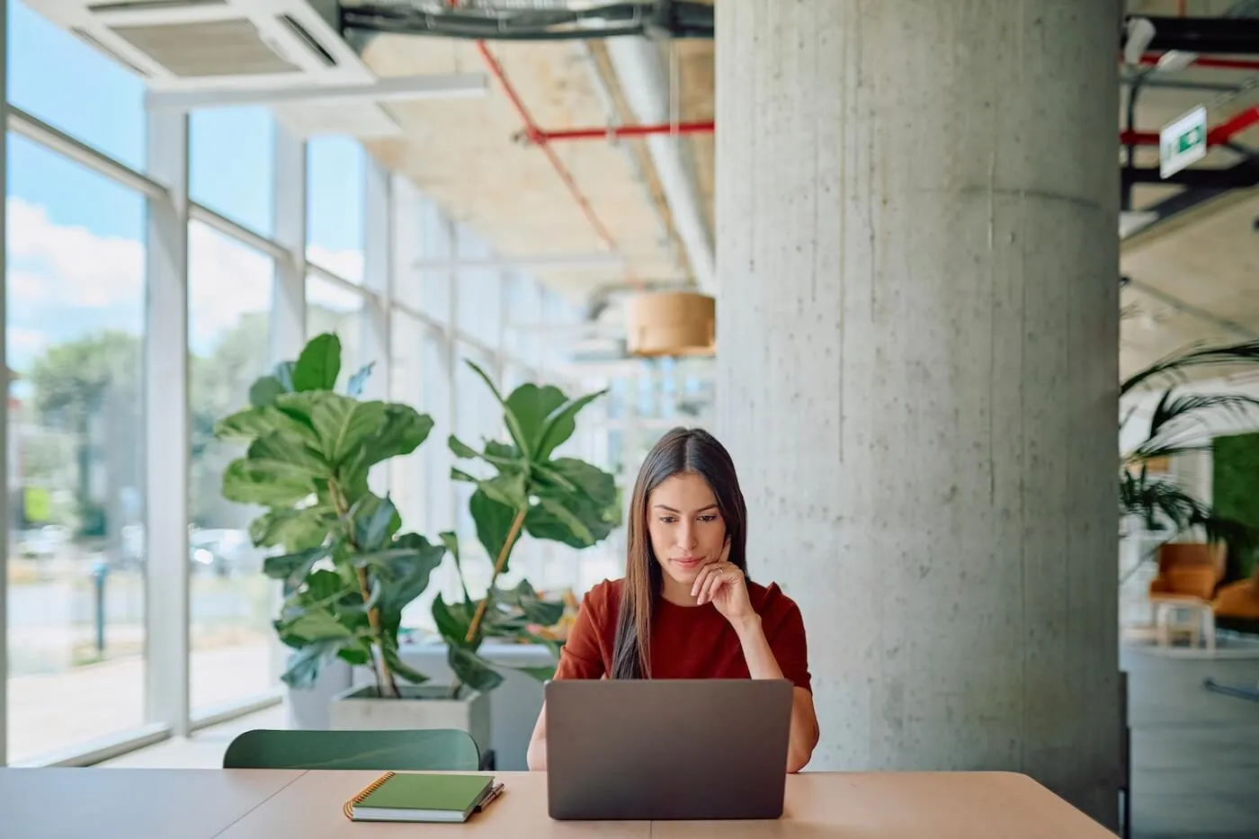 Focused young woman seated at a table using a laptop in a modern office workspace, with large windows, indoor plants, exposed concrete columns, and a notebook on the table nearby.