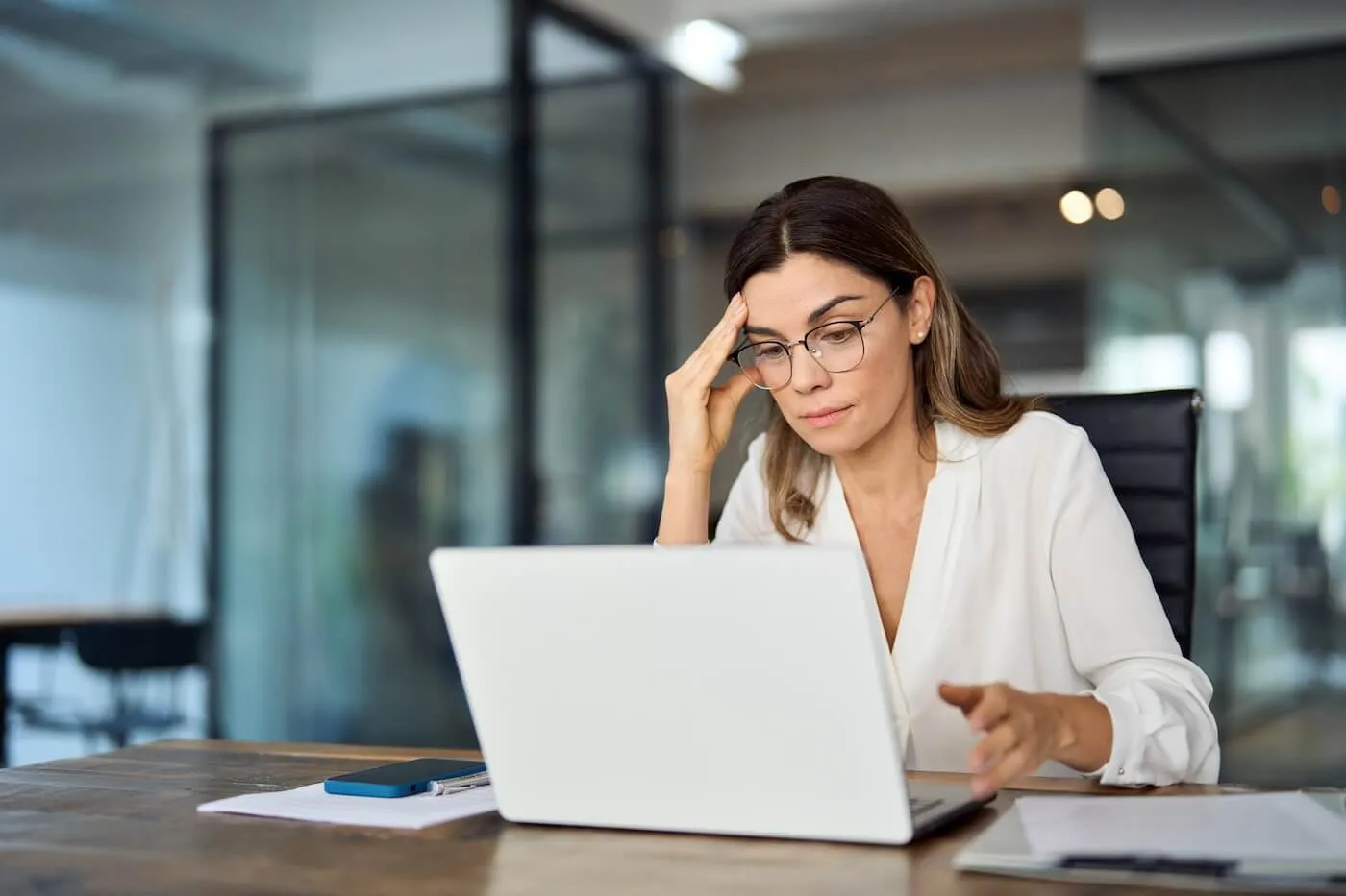 Focused woman wearing glasses working on a laptop at a desk in a modern office, with documents and a smartphone on the table and glass-walled meeting rooms in the background.