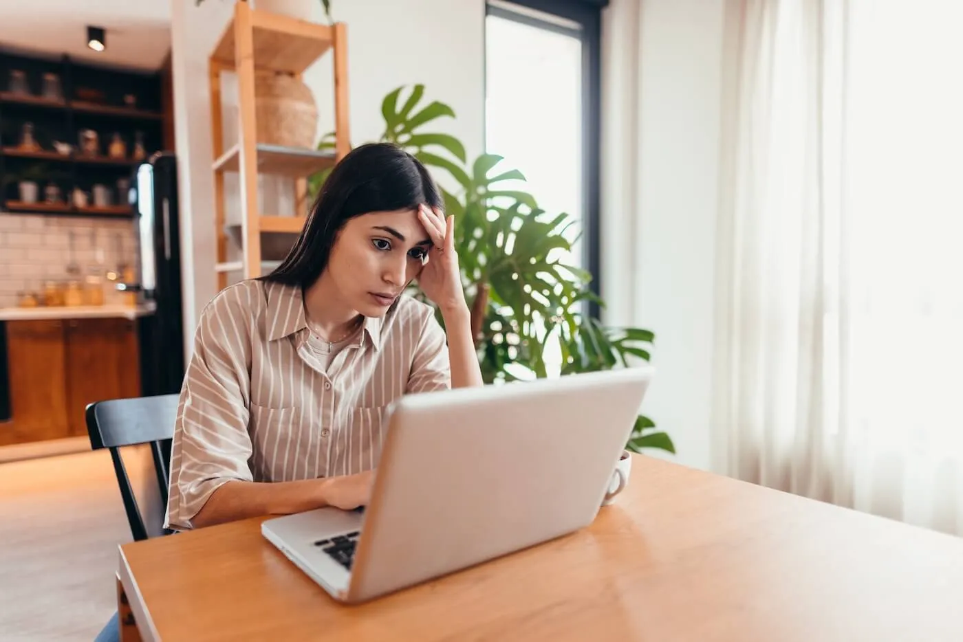 Concerned woman seated at a wooden table in a home setting, looking at a laptop with one hand resting against the forehead. A coffee cup sits beside the computer, while houseplants, shelving, and a bright window are visible in the background