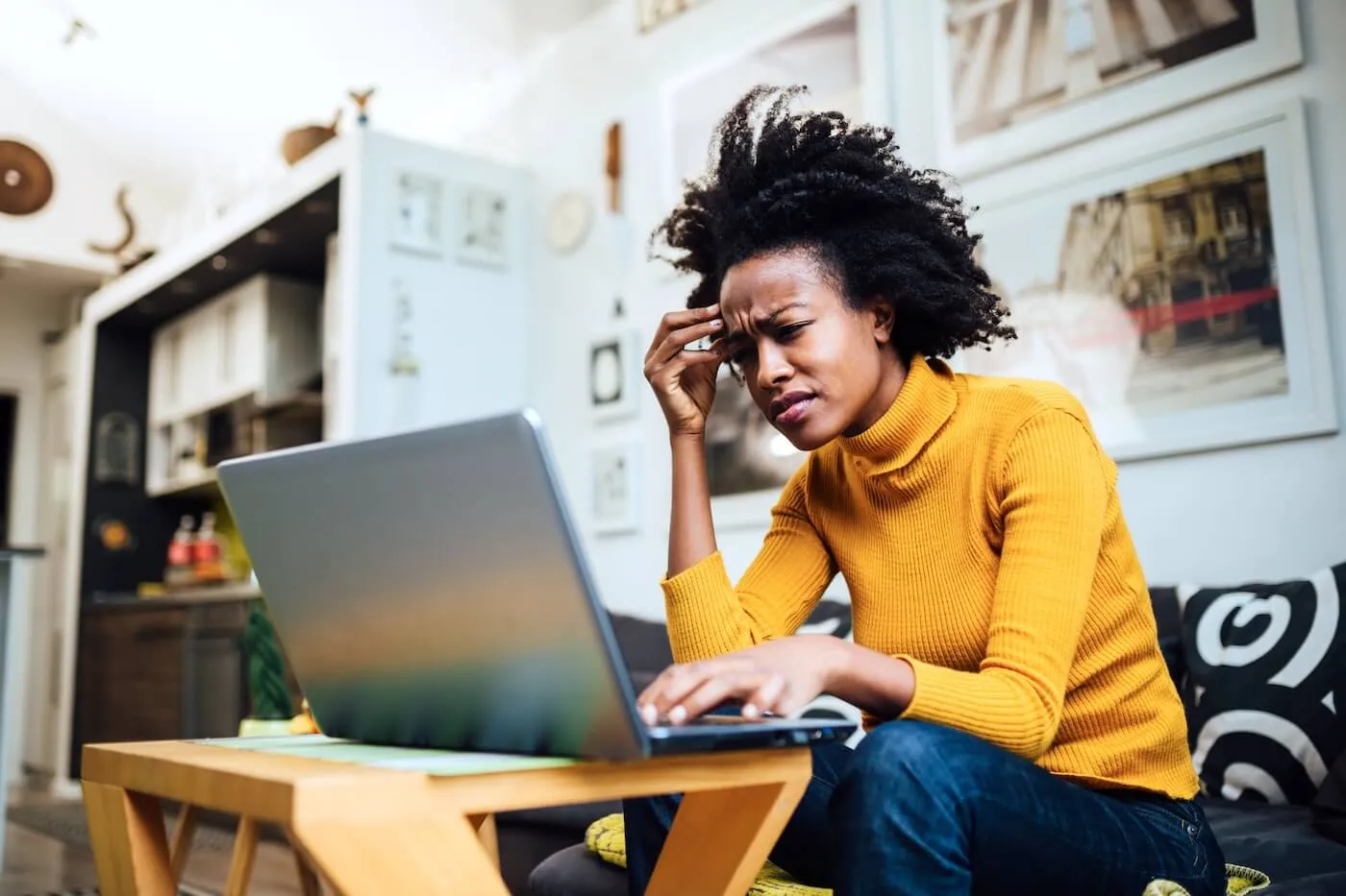 A worried woman seated on a sofa using a laptop on a coffee table in a home living room. Framed artwork, shelving, and home furnishings are visible in the background.