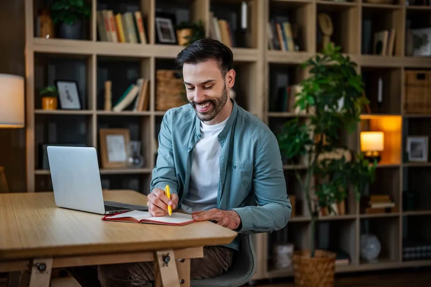 Smiling young man taking notes in a home office with an open laptop on a desk