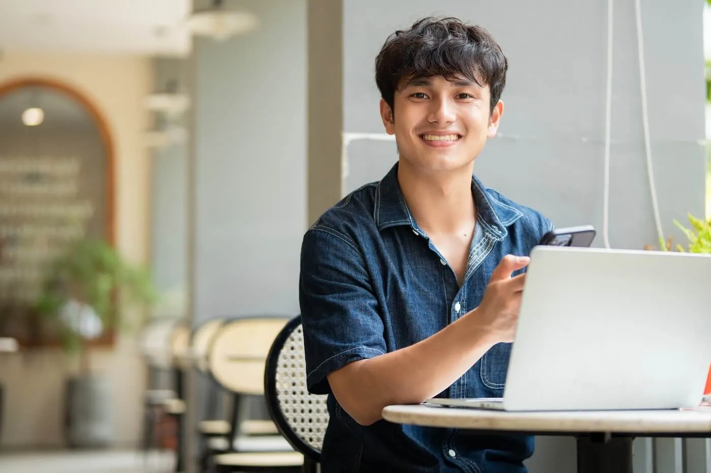 Smiling young man seated at a café table using a laptop while holding a smartphone, with indoor seating and natural light visible in the background.