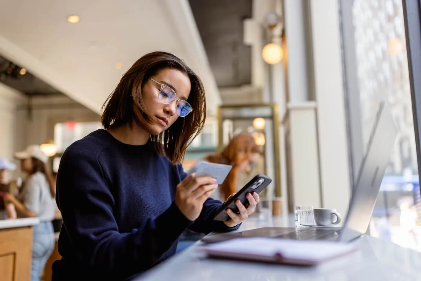Focused young woman sitting in the cafe, using her credit card and a smartphone, with an open laptop on the table