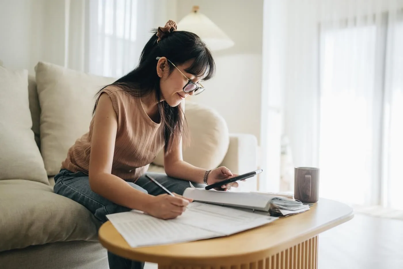 Focused young woman sitting on a sofa at home reviewing paperwork, writing notes, and using a smartphone at a coffee table with documents, a notebook, and a mug nearby.