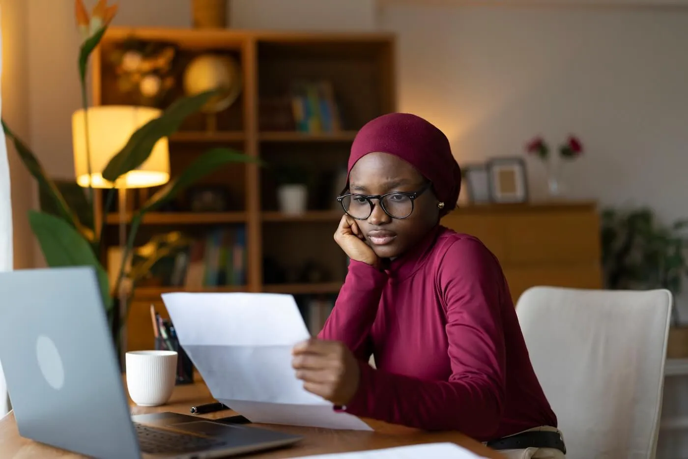 Focused young woman seated at a desk in a home office reviewing a printed document beside an open laptop. A coffee mug, pen, and papers are on the desk, with bookshelves, plants, and warm lighting visible in the background.