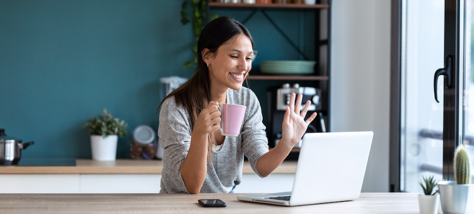 Smiling young woman waving through the laptop web camera while holding a cup of coffee in the kitchen at home.