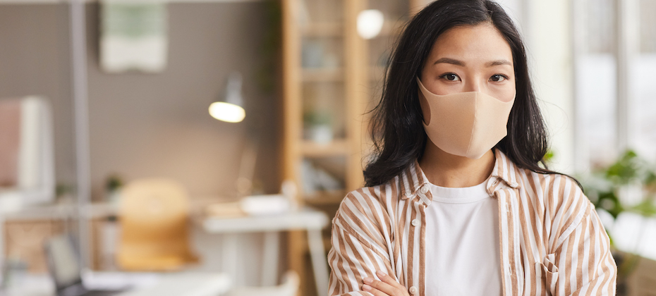 Young Asian woman wearing mask and looking a camera while standing with arms crossed in modern office