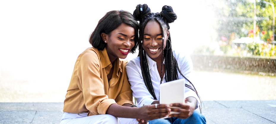 Two black girls sitting near fountain and looking video at digital tablet.