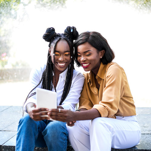 Two black women sitting near fountain and looking video at digital tablet.