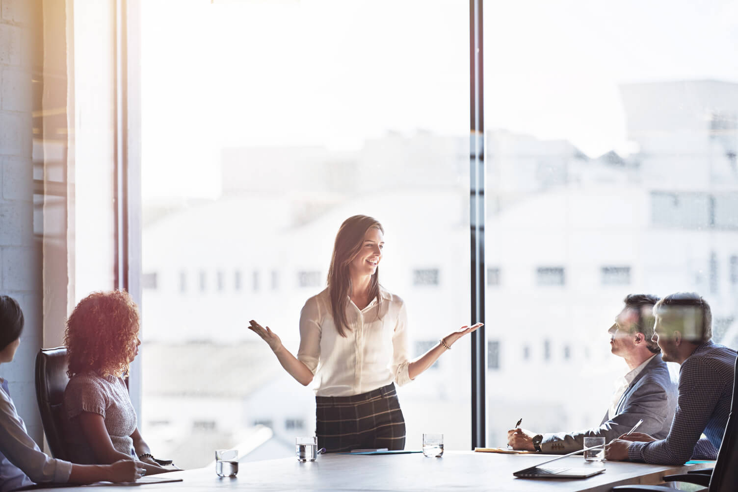 Woman presenting at a meeting to a group of seated colleagues in a bright office with city views.