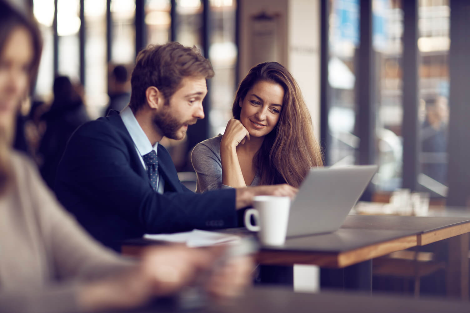 Two coworkers sit at a table in a bright café-like workspace, looking at a laptop together and smiling while discussing work. Other people are blurred in the background.
