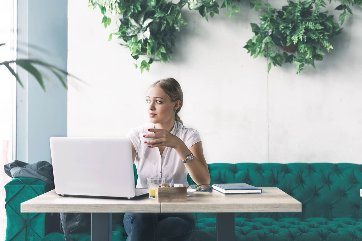 A woman in business casual attire works on a laptop at a table in a modern office lounge with green seating and wall plants.