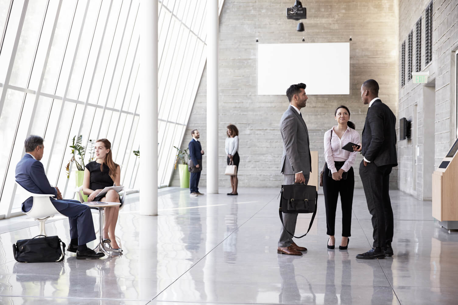 A bright, modern office lobby with floor-to-ceiling windows. Several small groups of professionally dressed people are talking or waiting. In the foreground, three colleagues stand in conversation, while another person sits with a laptop and another sits nearby. The space is open, polished, and business-like.