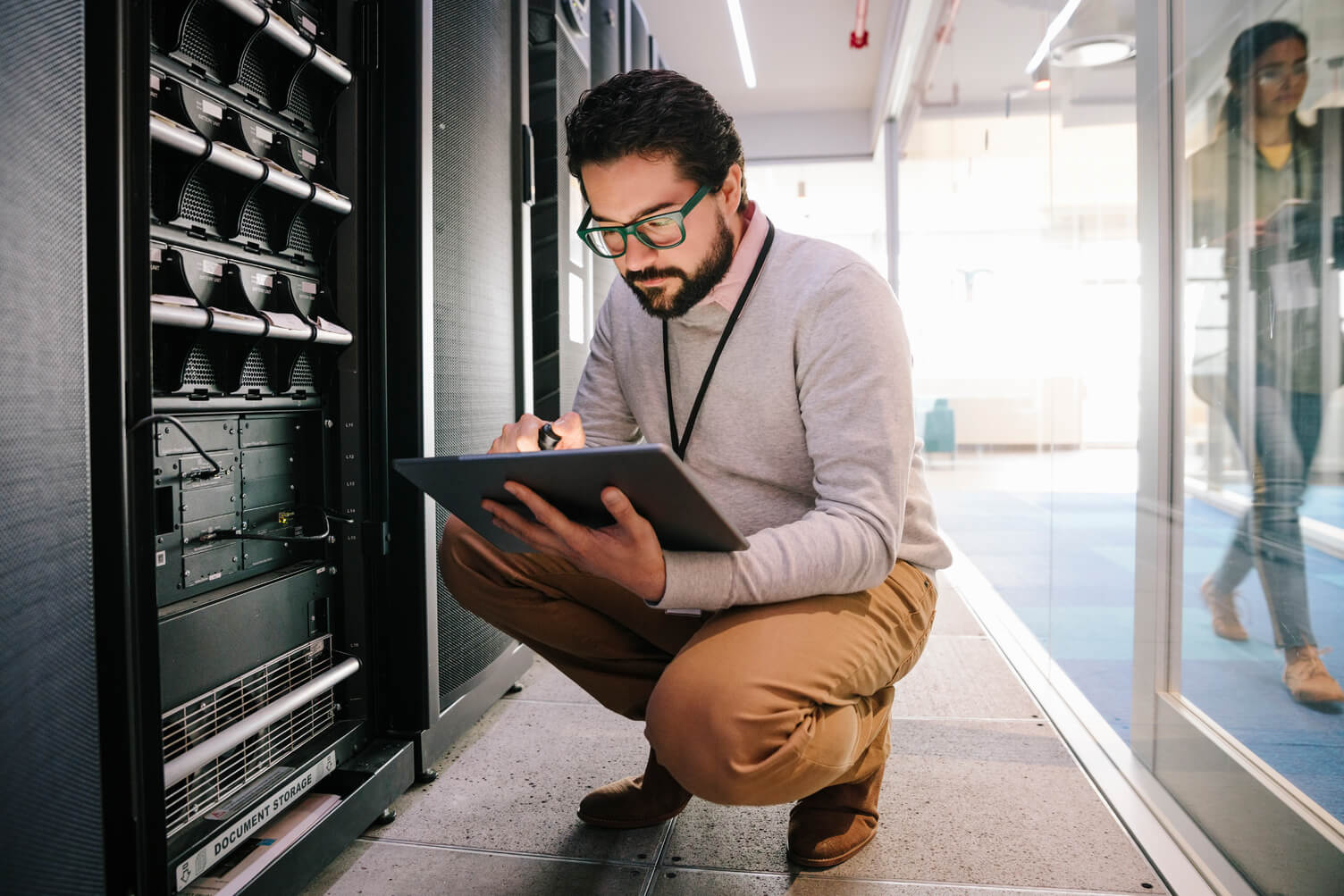 A man with glasses kneels beside a server rack, using a tablet in a modern data center hallway with glass office walls.