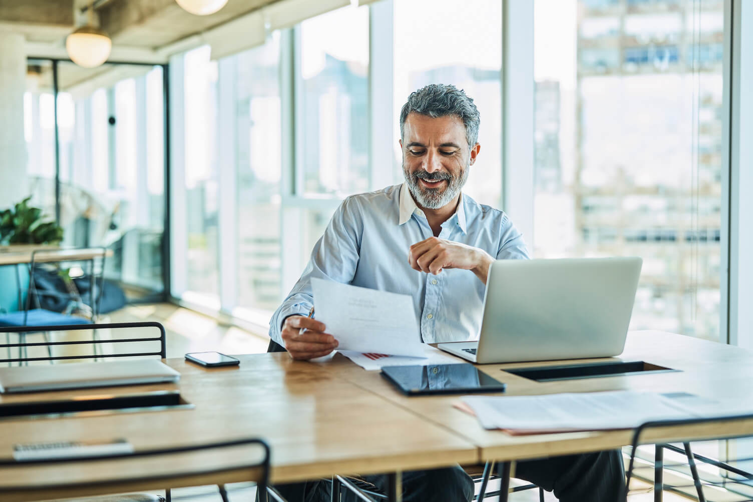 Businessman smiling while reading document. Confident male professional is at coworking space. He is sitting at table.