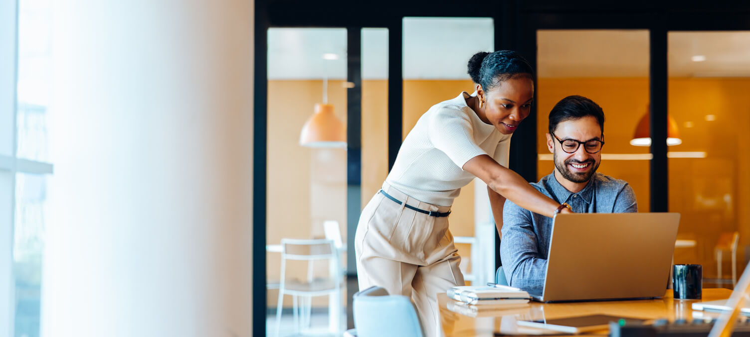 Two young professionals, a man and a woman, share ideas while reviewing work on a laptop in a corporate office setting, showcasing collaboration, productivity, and engagement at work.