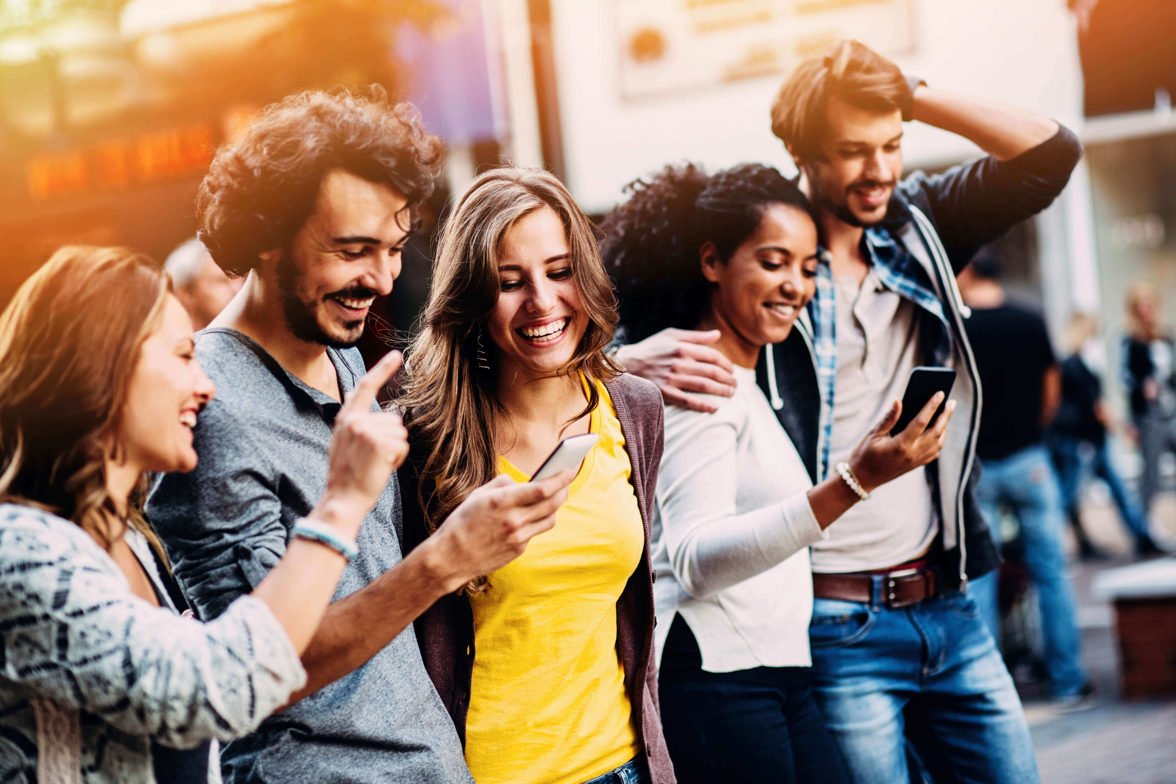 5 multi ethnic millenials cheerfully walking shoulder to shoulder in a line while looking at their phones.