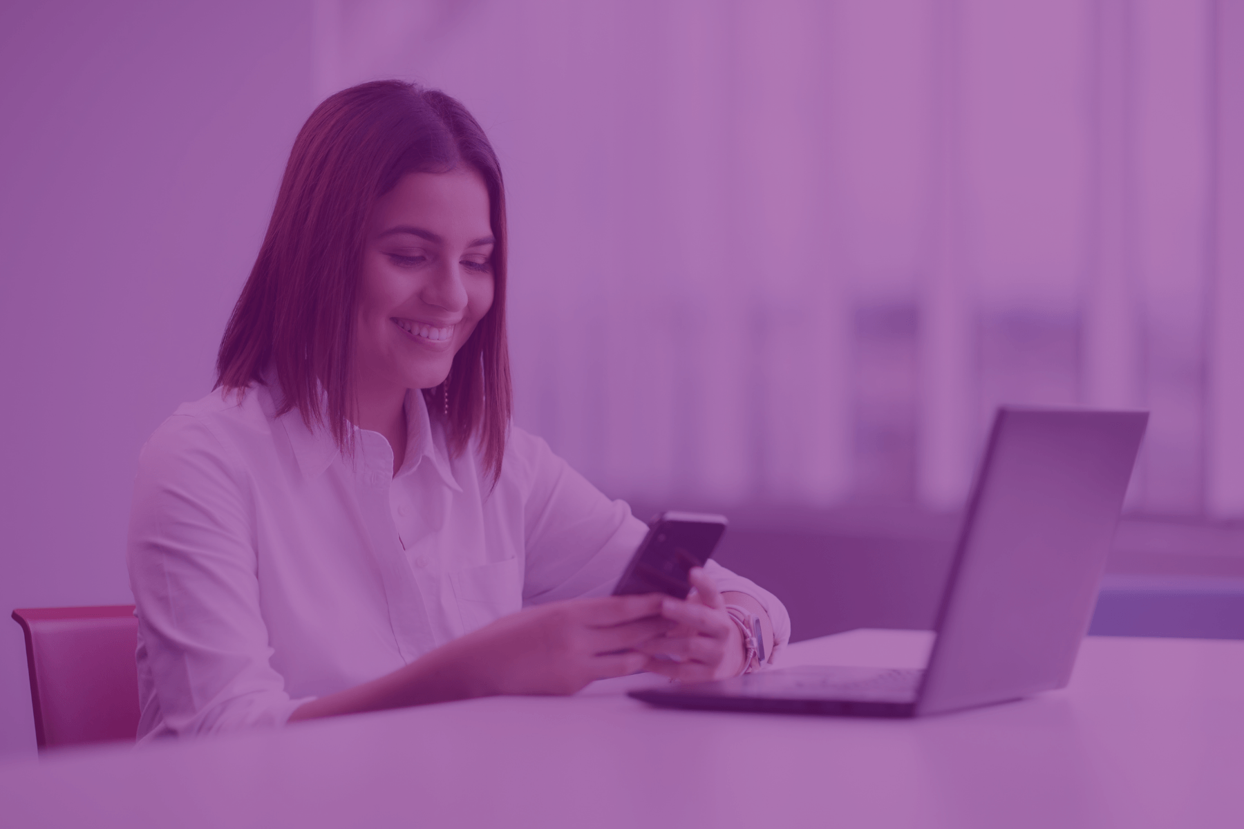 Smiling woman using a smartphone while working at a laptop.
