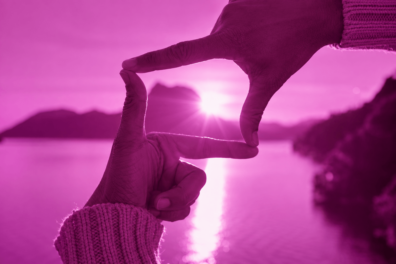 Two hands form a rectangular frame around the setting sun, overlooking a calm lake surrounded by dark mountain silhouettes, with warm sunlight reflecting across the water.