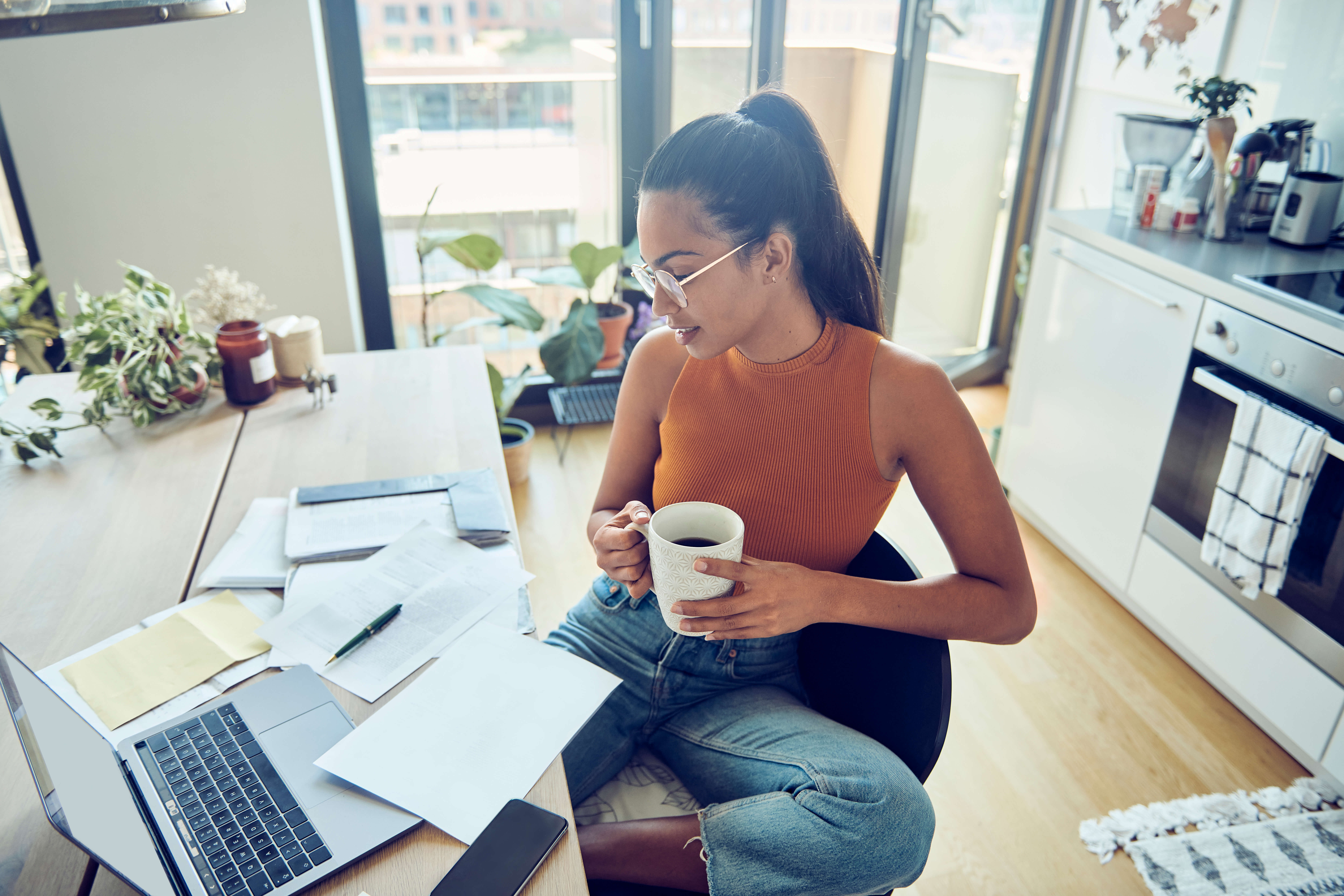 Woman with dark hair and glasses sits at a home office desk holding a coffee mug, with a laptop, paperwork, and bright kitchen space nearby.