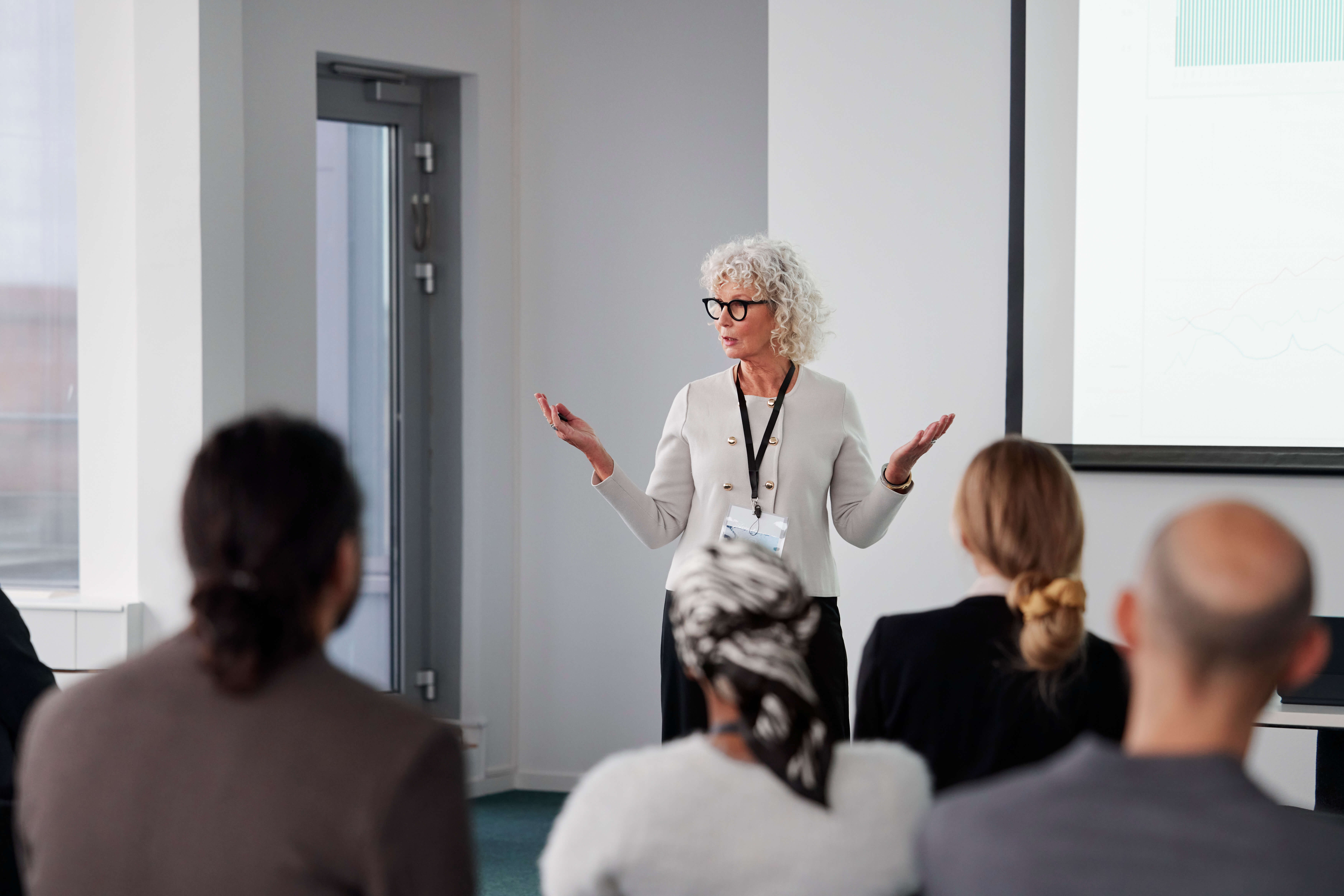 Older white woman with gray hair and glasses presents to a diverse group in a modern office training room, audience seated facing a screen.