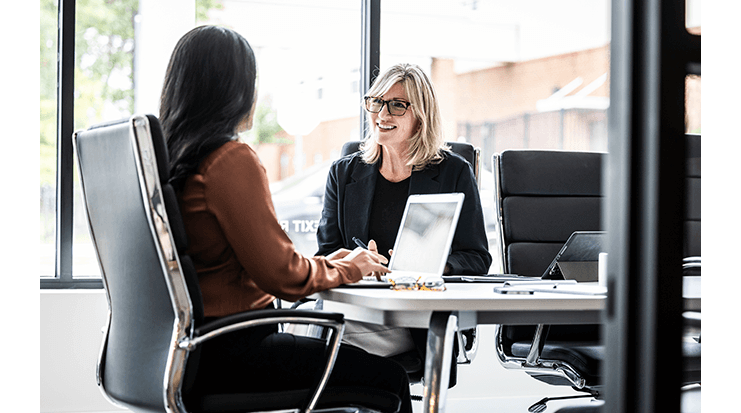 Black woman and white woman sit across a desk in a bright office, reviewing documents on a laptop during a verification meeting.