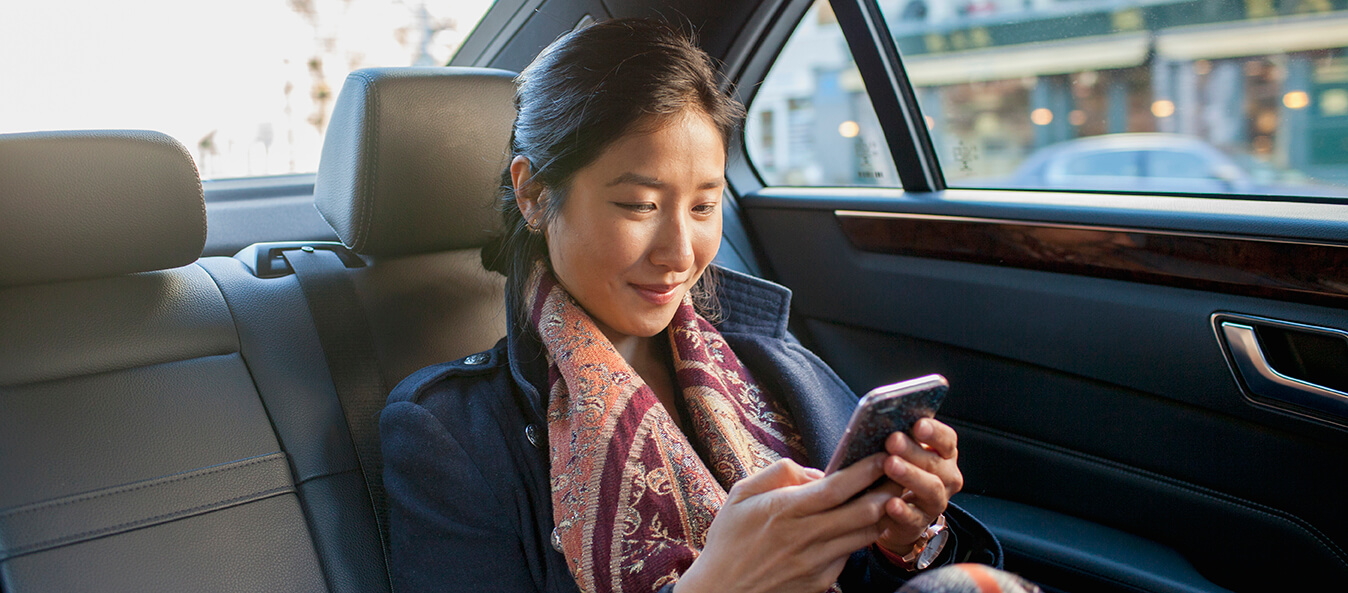 Young Asian woman cheerfully looking down at her phone in the back seat of a car.