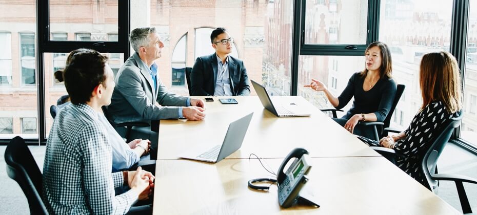 6 multi-ethnic business people sitting around a brightly lit board room talking.