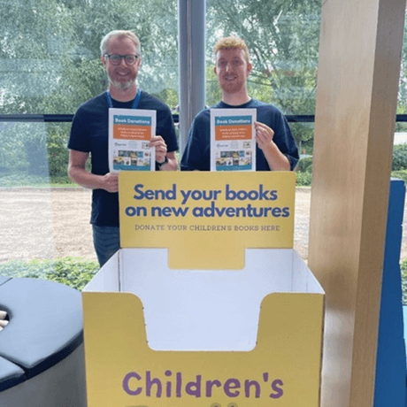 5 of 12 logos - Two people standing in front of a children's book drop box